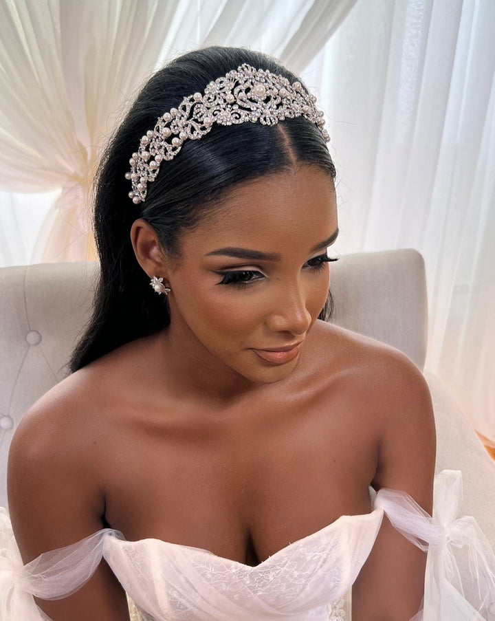 A woman sits gracefully in a white off-shoulder dress, her long dark hair cascading down. She wears a Vintage Inspired Swarovski Crystal Headband, style no. 80107, that glistens alongside her flower-shaped CZ wedding earrings earrings, while soft light through sheer curtains enhances her elegant aura.