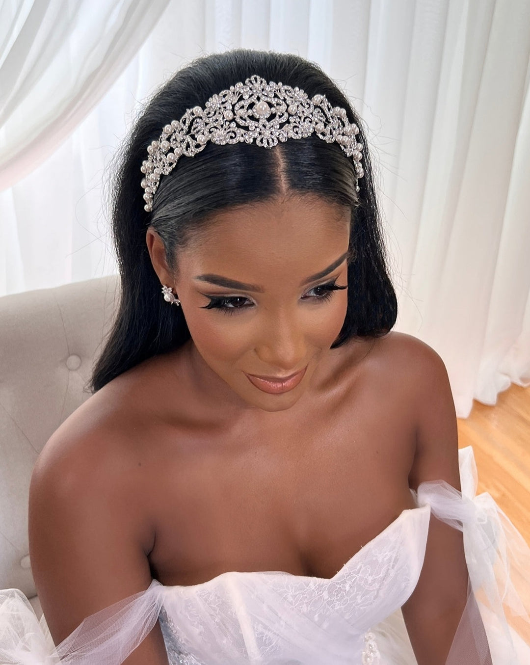 A woman with long dark hair, wearing a sparkling Vintage Inspired Swarovski Crystal Headband style no. 80107 and CZ wedding earrings, sits in a white bridal gown. She is indoors on a light-colored chair with sheer white curtains behind her.