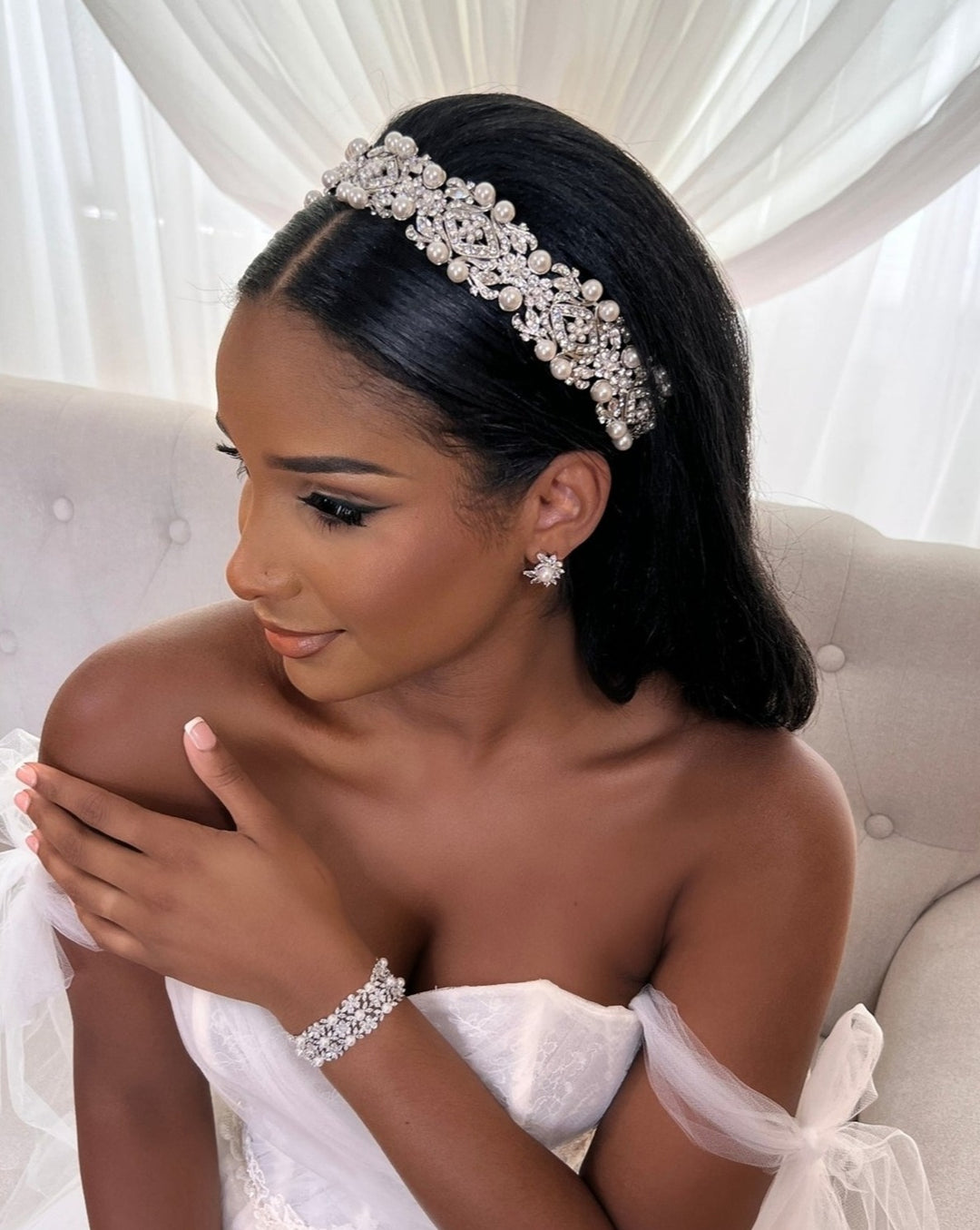 A woman with long dark hair wears a Vintage Inspired Swarovski Crystal Headband (style no. 80064), a strapless white dress, and matching wedding jewelry. She sits on a light-colored sofa with sheer curtains behind her, gazing to the left and gently touching her shoulder.
