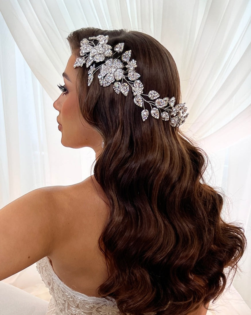 A woman with long, wavy brown hair wears the Crystal Flower & Leaflet Bridal Vine (Style no. 80014), a stunning Swarovski bridal hair accessory. She is in a strapless white gown, standing gracefully before sheer white curtains.