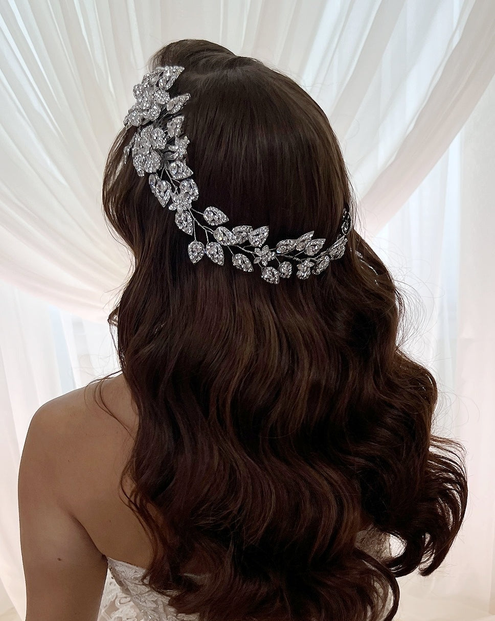 A woman with long, wavy brown hair and a Crystal Flower & Leaflet Bridal Vine (Style no. 80014) stands in front of sheer white curtains. She faces away from the camera, wearing a strapless white dress.