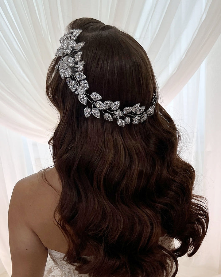 A woman with long, wavy brown hair and a Crystal Flower & Leaflet Bridal Vine (Style no. 80014) stands in front of sheer white curtains. She faces away from the camera, wearing a strapless white dress.