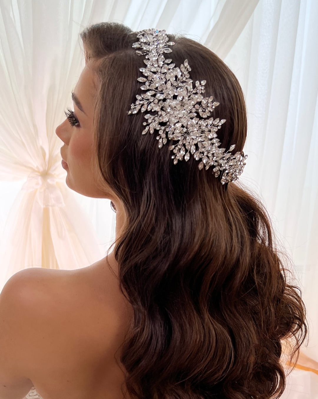 A woman with long, wavy brown hair is seen from behind, decorated with a Dramatic Swarovski  Crystal Bridal Halo style no. 242518, featuring intricate silver and crystal leaf designs. She stands before sheer white curtains as soft light filters through.