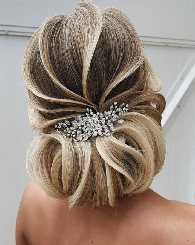 A woman with a chic updo of light brown and blonde highlights wears her hair in a looped pattern, decorated with the Dainty Crystal Bridal Comb (style no. 242731), which features a floral design. The background is light and neutral.