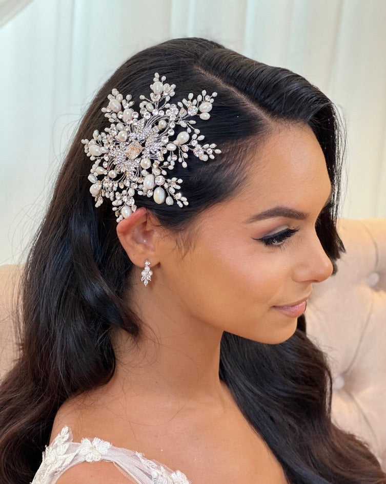 A woman with long dark hair wears the Dreamy Pearl and Crystal Comb (style no. 3973670), featuring Swarovski crystals and pearls, along with matching earrings. She is dressed in a white lace outfit, seated against a light-colored background.