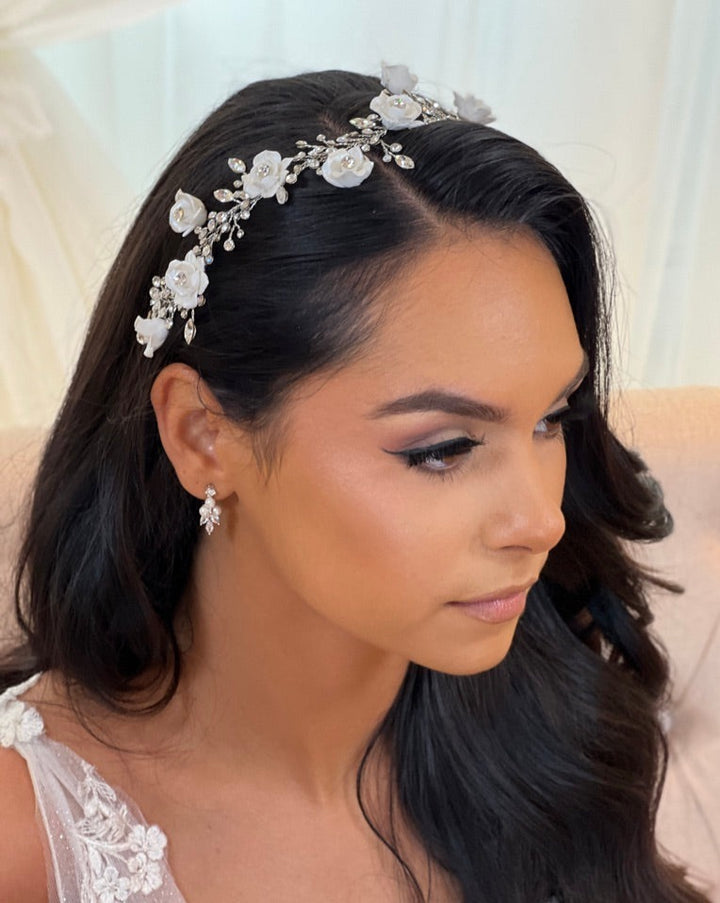 A woman with long dark hair wears a  Romantic Flower Swarovski Hair Vine style no. 242720 with white flowers and crystals. She has subtle makeup, a lace-strap white dress, and is seated in a softly lit room.