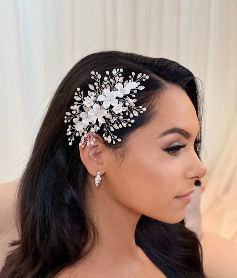 A woman with long, dark hair wears the Porcelain Flower Bouquet Comb (style 242722) studded with Swarovski crystals and pearls. She has subtle makeup and delicate, dangling earrings. Her gaze is turned to the side against a light background.