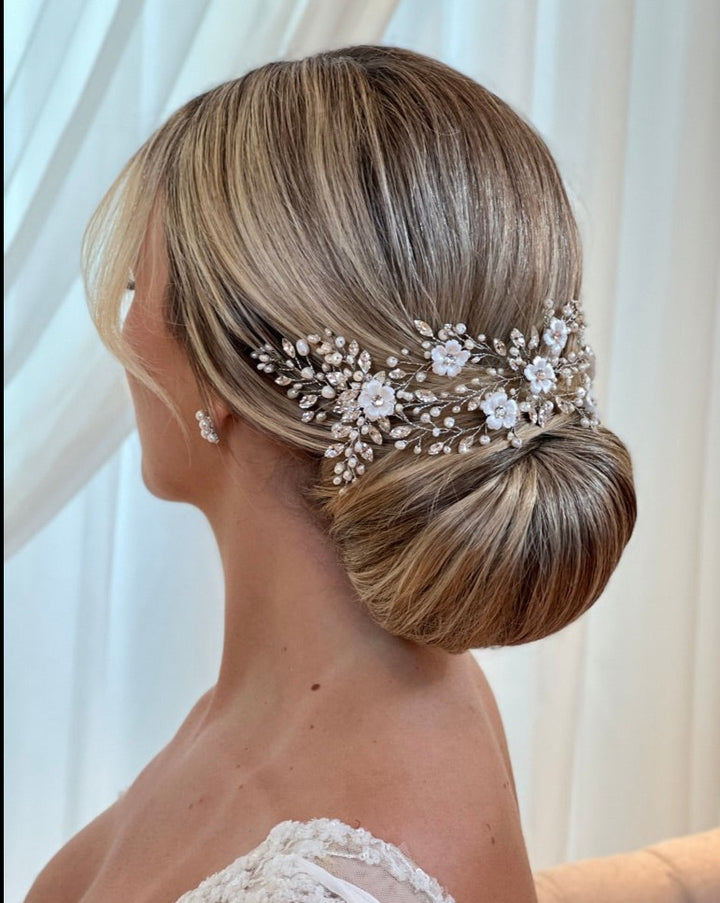 A woman with blonde hair in a low bun wears the Delicate Pearl, Porcelain, and Swarovski Flower Bridal Hair Vine style no. 242712 complemented by small bridal earrings and a white lace dress. She stands against a softly lit curtain backdrop.