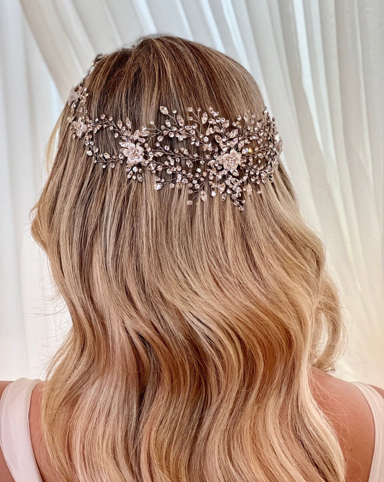 A person with long, wavy blonde hair wears the Enchanting Crystal and Flower Wedding Halo, style no. 242713, adorned with intricate floral designs and sparkling Swarovski crystal embellishments, against a softly lit, light-colored background.