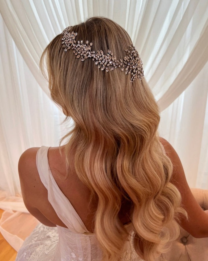 A woman with long, wavy blonde hair faces away, adorned with the intricate Dramatic Crystal Halo style no. 242518 Swarovski wedding headpiece. Shes wearing an elegant white dress against a backdrop of soft, flowing white curtains.