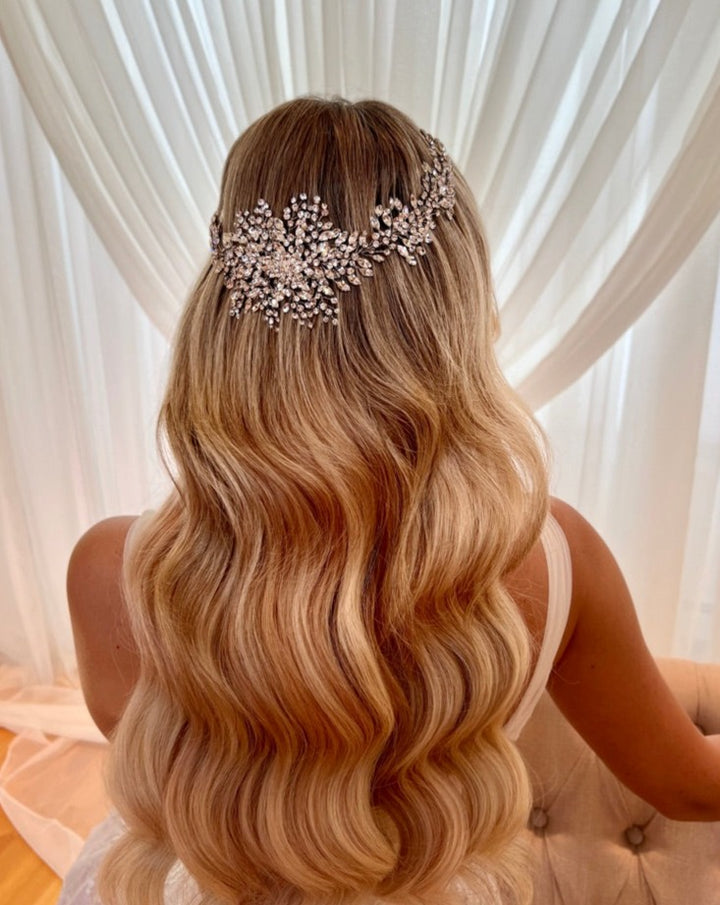 A woman with long, wavy blonde hair faces away from the camera, wearing a Dramatic Swarovski  Crystal Bridal  Halo crown (style no. 242518). Sheer white drapes in the background create a soft, elegant setting.